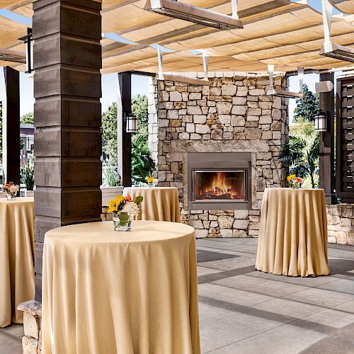 An outdoor event space with tall, round tables covered in beige tablecloths, small floral centerpieces, a stone fireplace, and a shaded canopy.