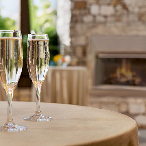 Two champagne flutes on a table with a flower arrangement nearby, and a fireplace in the background within a cozy setting.