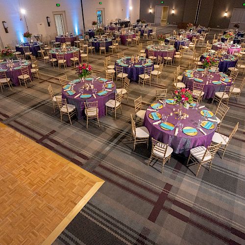 A decorated banquet hall with round tables set with purple tablecloths, arranged place settings, floral centerpieces, and chairs around them.