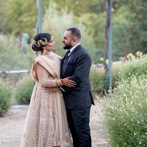 A man and a woman, dressed in formal attire, are gazing at each other lovingly while standing in a garden setting.