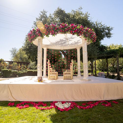 An outdoor wedding mandap decorated with red flowers and drapery, set on a platform with two wooden chairs under a white canopy.