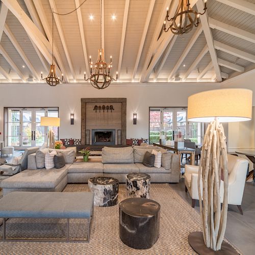 A spacious, modern living room with high ceilings, chandeliers, soft seating, rustic tables, and a contemporary fireplace, creating a cozy ambiance.