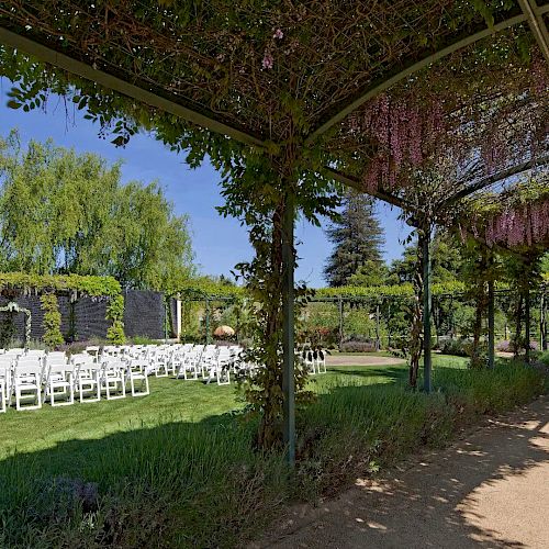 The image shows an outdoor garden setup with white chairs arranged for a ceremony, possibly a wedding. A pathway with lush greenery is also visible.