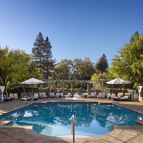 A serene outdoor pool surrounded by lounge chairs, umbrellas, and lush greenery under clear blue skies in a tranquil setting.