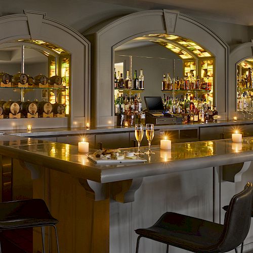 A dimly lit bar with candles, bottles of liquor, mirrors, a bar counter, chairs, and glasses of champagne on the counter.