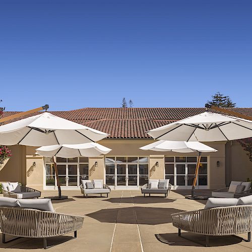 The image shows an outdoor courtyard with beige buildings, large white umbrellas, and comfortable seating arranged around tables.