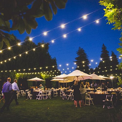 An outdoor event with string lights, guests mingling, round tables under umbrellas, and a scenic evening backdrop in a garden setting.