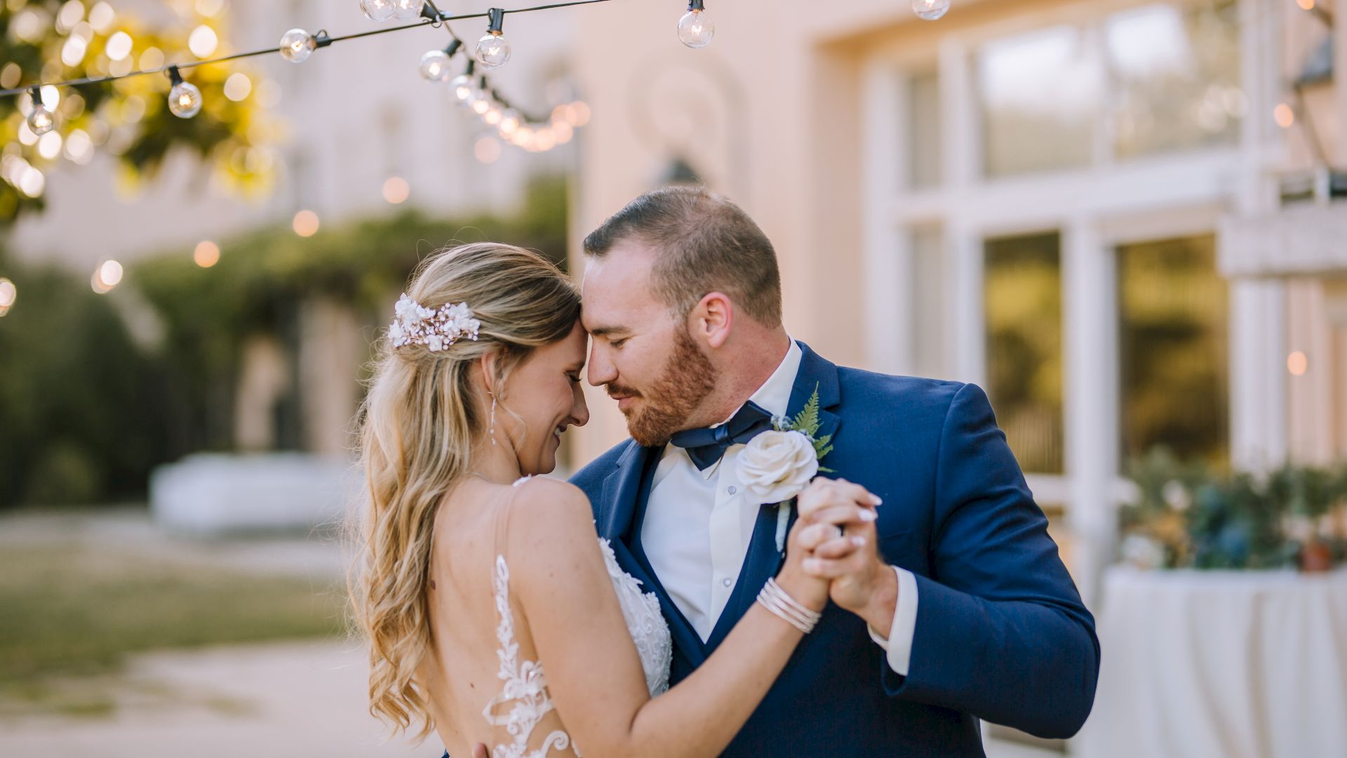 A bride and groom share a romantic dance under string lights outdoors, with the groom in a blue suit and the bride in a lace dress.