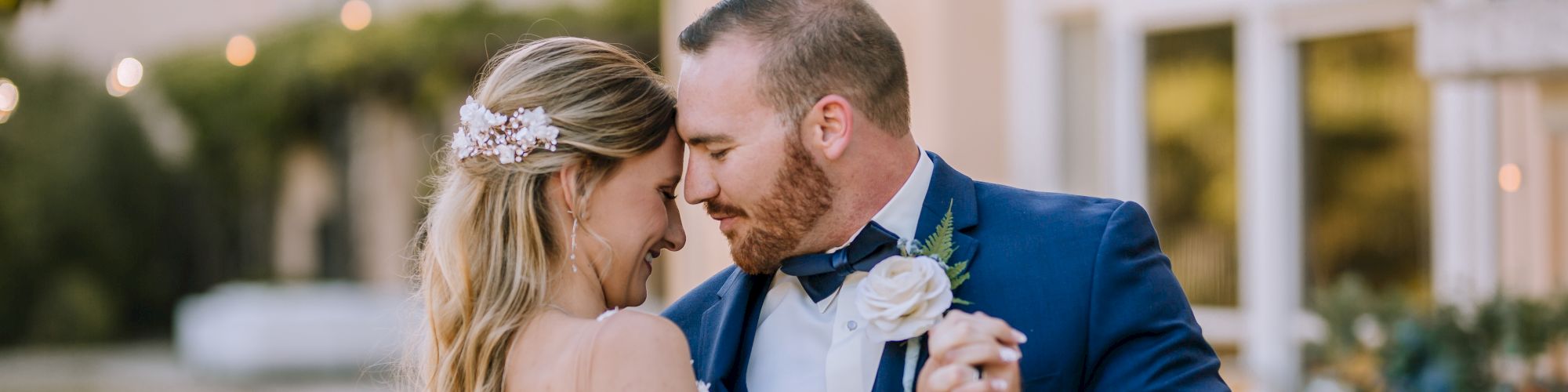 A bride and groom share a romantic dance under string lights outdoors, with the groom in a blue suit and the bride in a lace dress.