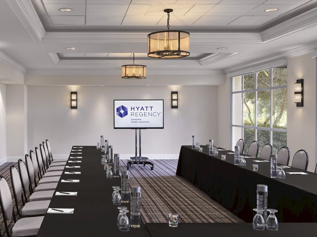 A conference room with a U-shaped table setup, black tablecloths, water pitchers, glasses, chairs, a screen displaying "Hyatt Regency," and large windows.