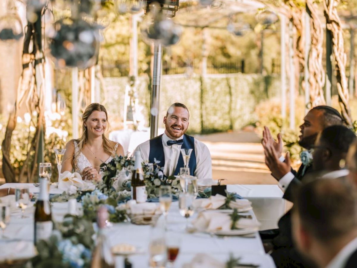A smiling bride and groom are seated at a beautifully decorated table, surrounded by guests, likely during a wedding reception outdoors.