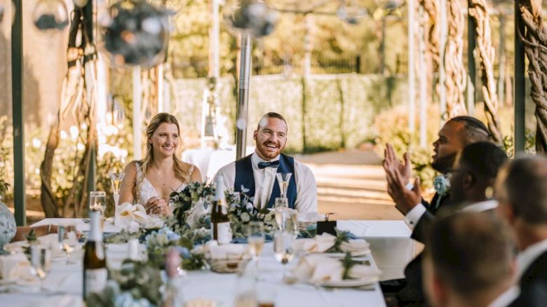 A smiling bride and groom are seated at a beautifully decorated table, surrounded by guests, likely during a wedding reception outdoors.