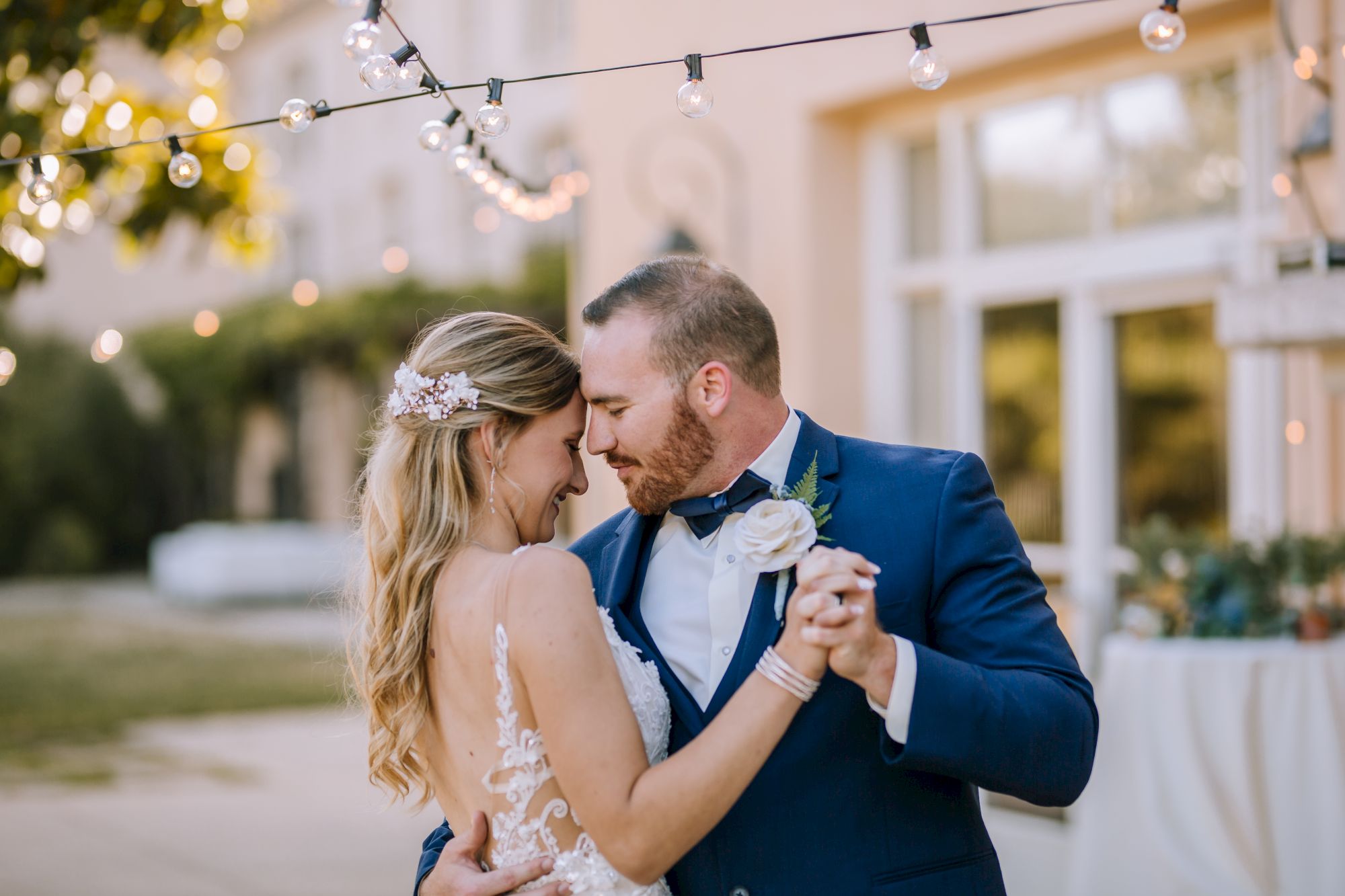 A couple dressed in wedding attire shares a romantic dance outdoors under string lights, with greenery and a building in the background.