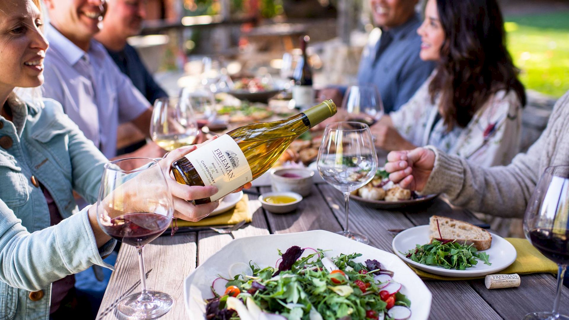 A group of people enjoying a meal outdoors, with wine being poured and a table filled with fresh salads, bread, and other dishes.