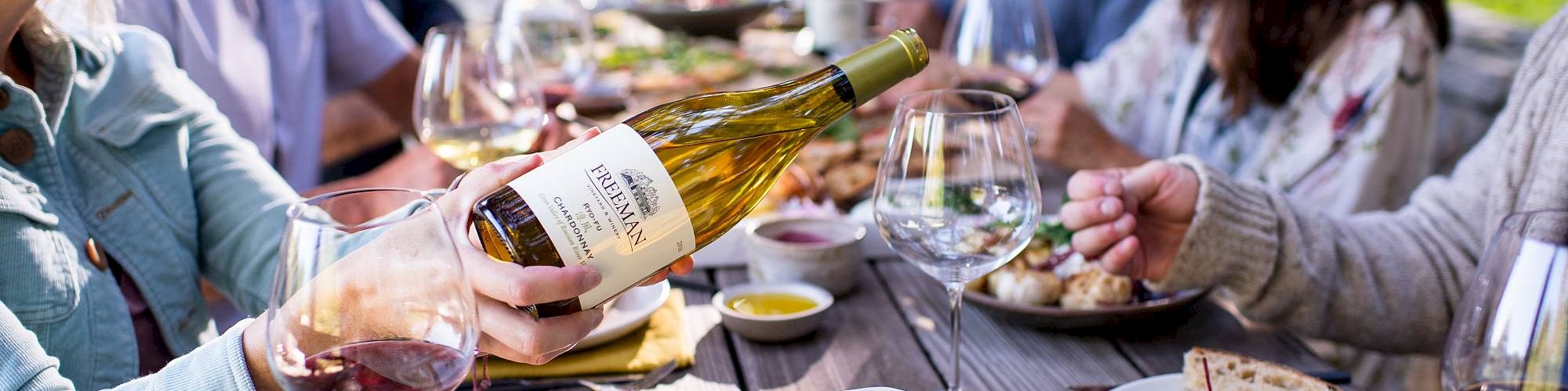 A group of people enjoying a meal outdoors, with wine being poured and a table filled with fresh salads, bread, and other dishes.