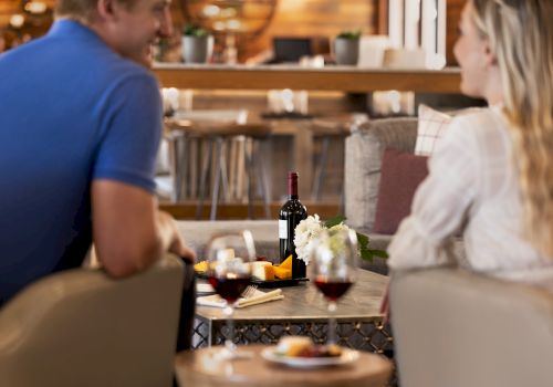 A couple is seated at a table with wine and cheese, engaging in conversation in a cozy, well-decorated setting.