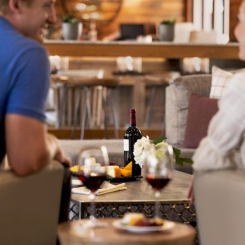 A couple is seated at a table with wine and cheese, engaging in conversation in a cozy, well-decorated setting.