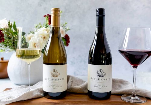 Two bottles of MacRostie wine, a white and a red, are displayed alongside glasses of wine and a floral arrangement on a wooden surface.