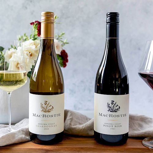 Two bottles of MacRostie wine, a white and a red, are displayed alongside glasses of wine and a floral arrangement on a wooden surface.