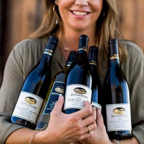 A person is holding several bottles of wine, smiling at the camera. The wine labels read "Dutton Estate" and indicate different varieties.