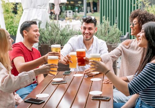 A group of five people are sitting outdoors around a wooden table, smiling and raising glasses, appearing to be enjoying drinks together.