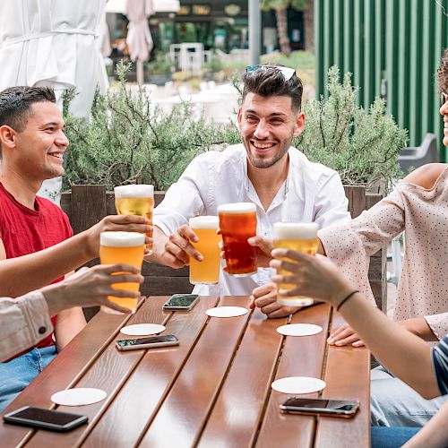 A group of five people are sitting outdoors around a wooden table, smiling and raising glasses, appearing to be enjoying drinks together.