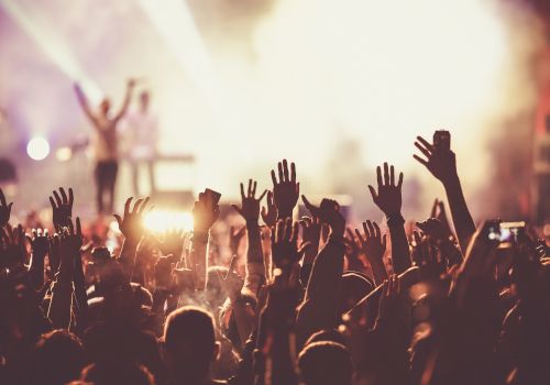 A lively concert scene with a crowd of people raising their hands, a brightly lit stage, and performers in the background.