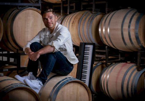 A man sits on a wine barrel in a cellar surrounded by more barrels with a keyboard leaning against them.