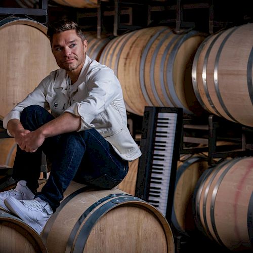 A man sits on a wine barrel in a cellar surrounded by more barrels with a keyboard leaning against them.