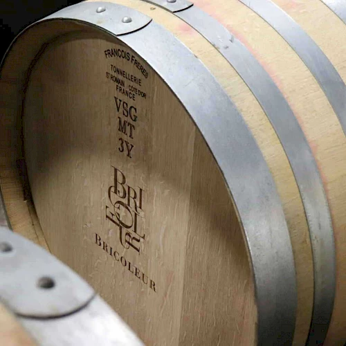 Wooden wine barrels with metal hoops and text indicating "VSG MT 3Y" and "BRI Bacqueyr" in black, likely in a wine cellar for aging.