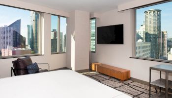 A modern hotel room with city views out large windows, a bed, chair, wall-mounted TV, and a wooden bench underneath the TV.