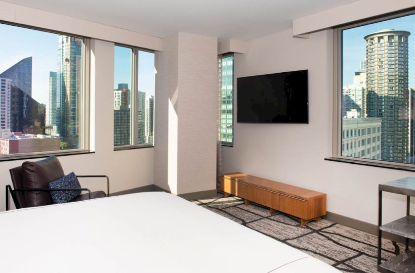 A modern hotel room with city views out large windows, a bed, chair, wall-mounted TV, and a wooden bench underneath the TV.