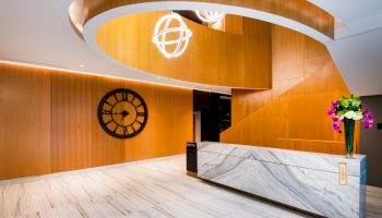 The image shows a modern interior with a marble reception desk, wooden walls, a large wall clock, and elegant lighting fixtures hanging from the ceiling.