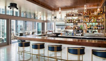A modern bar with a well-stocked liquor display, stylish seating, and large windows overlooking an urban cityscape, appears ready for patrons.