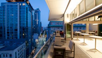 A modern balcony overlooking a cityscape at dusk, with seating areas and retractable glass walls, set against a backdrop of tall buildings.