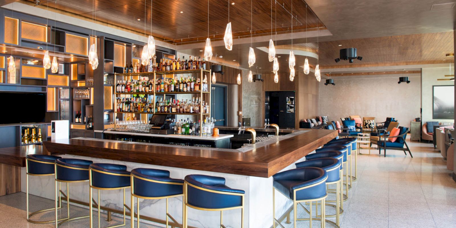 The image shows a modern bar with a sleek counter, stylish barstools, hanging lights, shelves stocked with various bottles, and a seating area in the background.