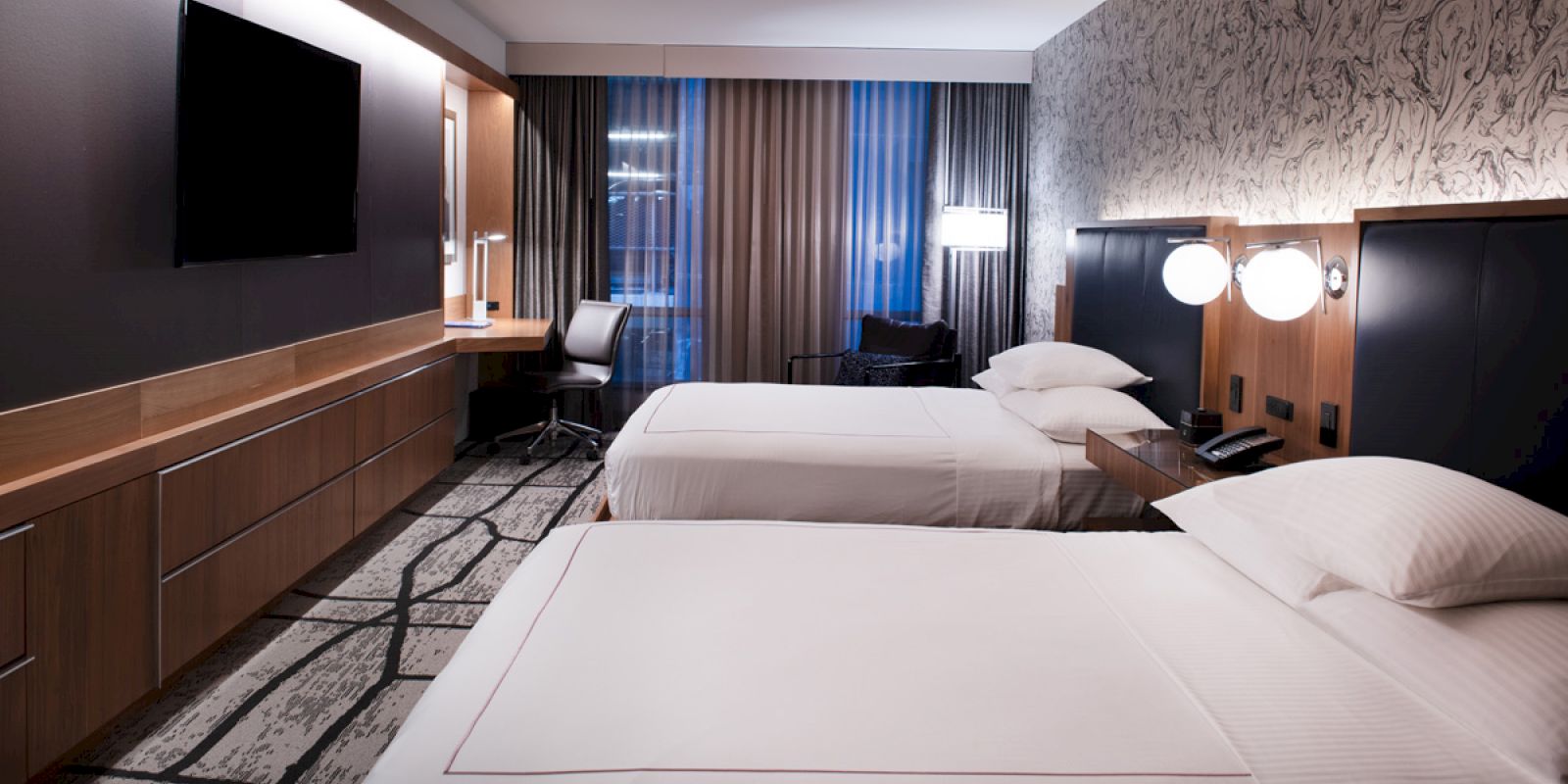 A hotel room with two neatly made beds, a wall-mounted TV, a desk with a chair, modern lighting, and patterned wallpaper and curtains.