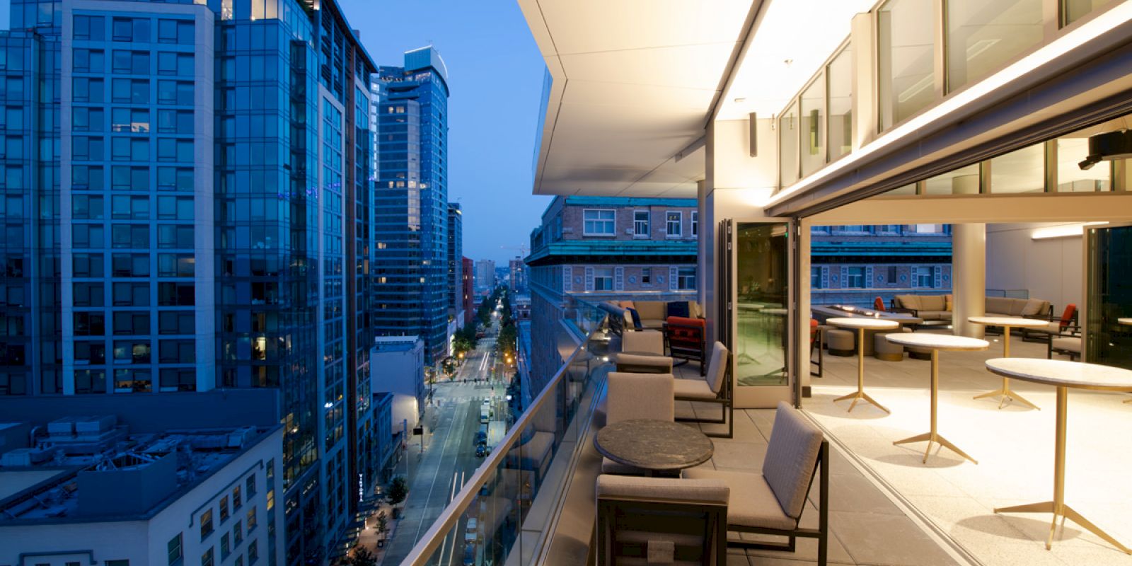 A modern cityscape view from a high-rise terrace at dusk, featuring outdoor seating, tables, and adjacent glass buildings in the distance.