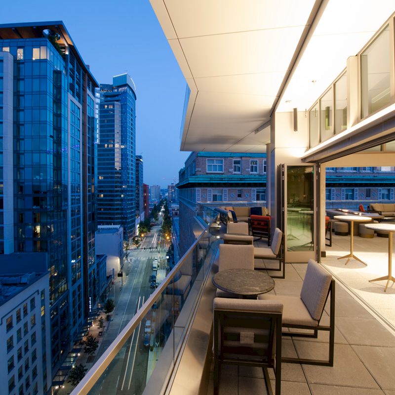 A modern cityscape with high-rise buildings is seen from a balcony, which is furnished with tables and chairs, during twilight.