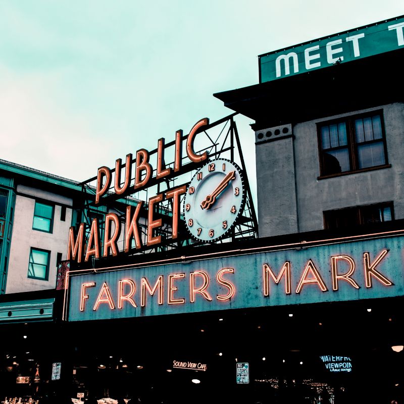 This image shows the iconic neon sign for a public market and a farmers market, along with a large clock on a building in an urban setting.