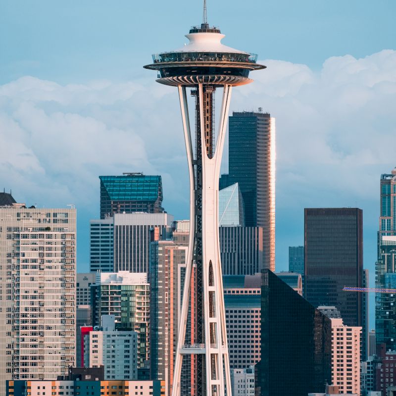 The image shows the Space Needle with a backdrop of skyscrapers and buildings under a cloudy sky, representing a bustling urban skyline.