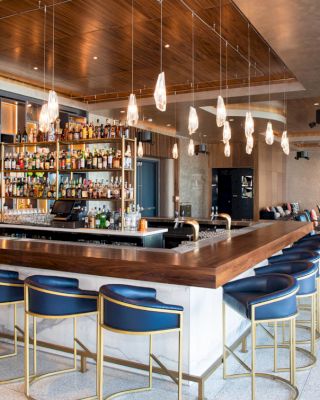 A modern bar with a wooden countertop and blue stools, hanging lights, and a well-stocked shelf of drinks in a spacious, elegant setting.