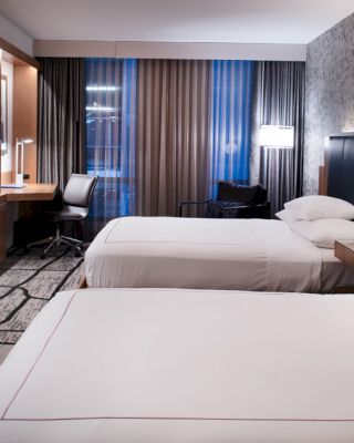 A modern hotel room with two double beds, a wall-mounted TV, a desk, a chair, and stylish lighting and decor for a comfortable stay.