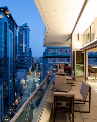 A rooftop balcony with tables and chairs overlooks a cityscape with tall buildings during early evening.