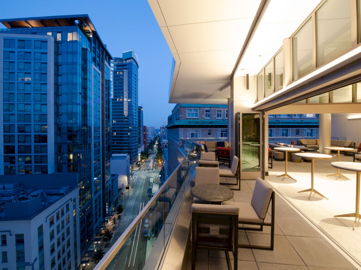A modern balcony with seating overlooks a city street lined with tall buildings at dusk, featuring a cozy, illuminated indoor space to the right.
