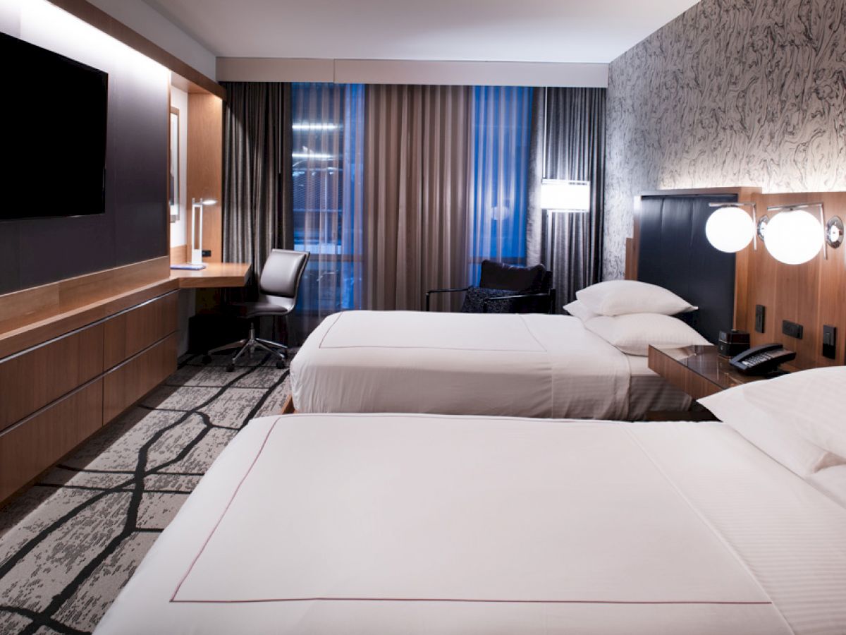 The image shows a modern hotel room with two neatly made beds, a wall-mounted TV, a desk with a chair, and stylish decor.