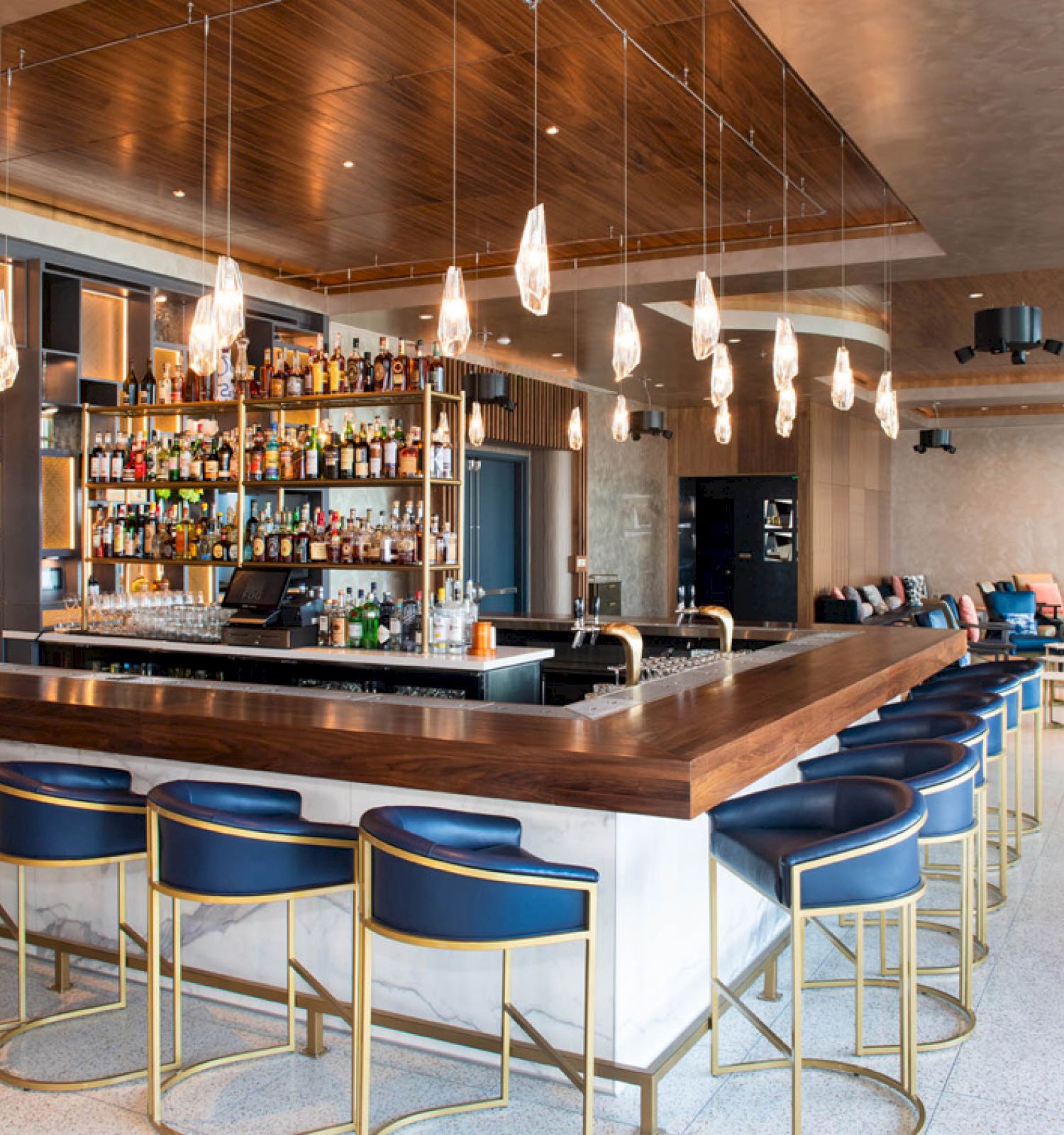 A modern bar with a wooden countertop, blue barstools, hanging lights, shelves with bottles, and seating areas in the background.