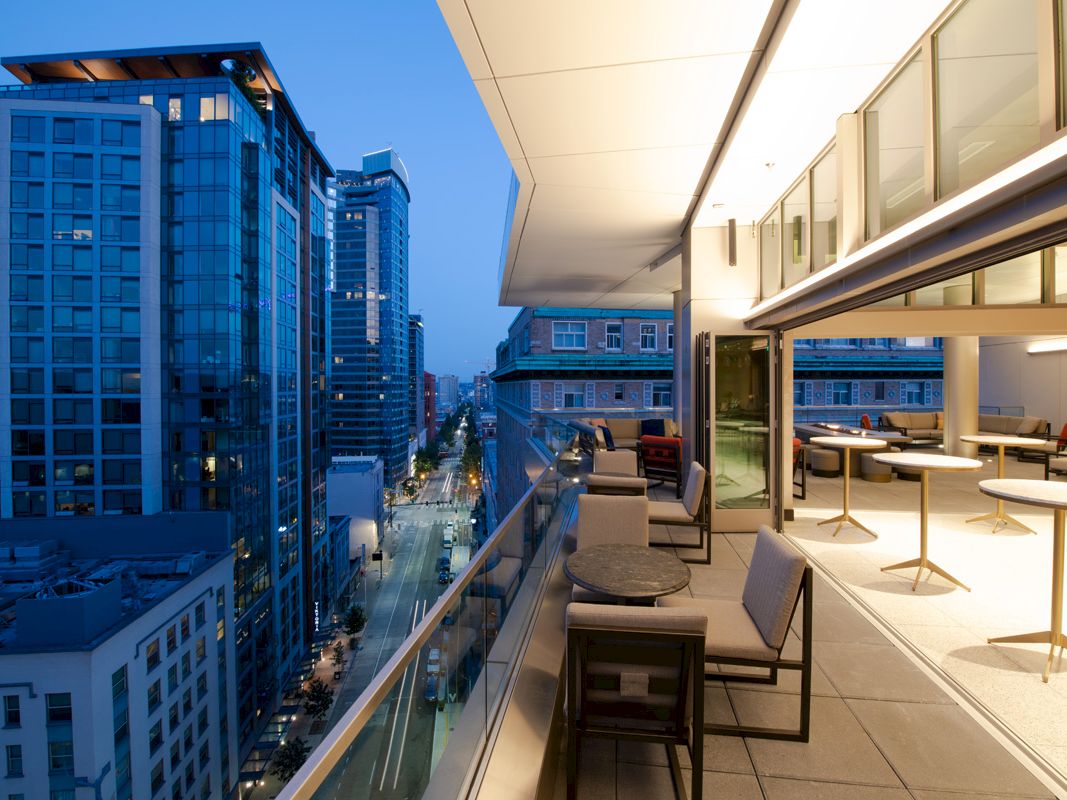 This image shows a modern urban balcony at twilight with outdoor seating, overlooking a cityscape with tall buildings and street lights below.