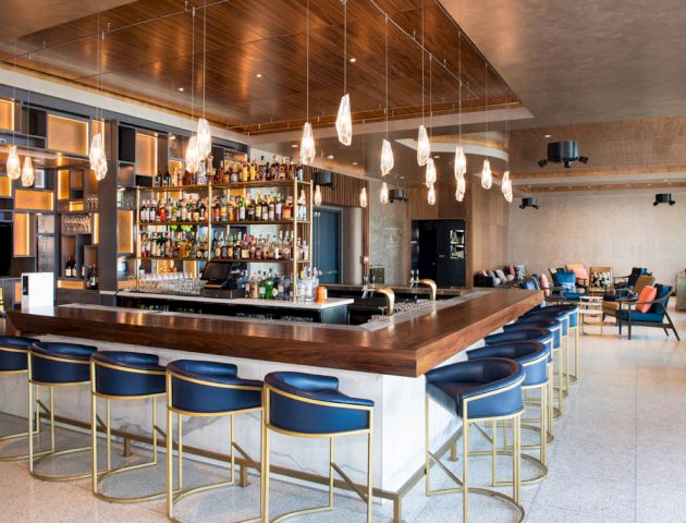A modern bar with blue bar stools, a wooden counter, pendant lights, and a well-stocked shelf of liquor bottles, set in a stylish, spacious room.