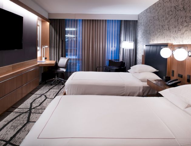 A modern hotel room with two beds, a flat-screen TV, a desk, and a window with curtains. The room features contemporary furnishings and lighting.